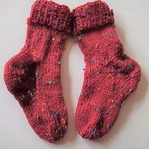 Könnte beinhalten: Ein Paar rote Stricksocken mit einem gesprenkelten Muster. Die Socken haben einen umgeschlagenen Bund und sind aus weichem Garn gefertigt.