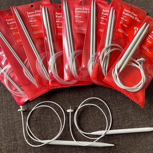 Könnte beinhalten: Eine Sammlung von Aero-Flex Stricknadeln in roter Verpackung. Die Nadeln sind in verschiedenen Größen erhältlich, darunter 2 mm, 3 mm, 4 mm, 5 mm und 7 mm, und sind 60 cm lang. Das Bild zeigt auch lose Stricknadeln.