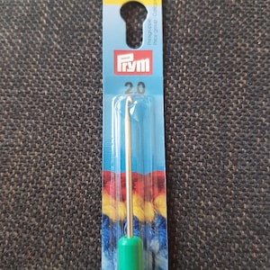 May include: A green crochet hook with a metal hook and a blue and yellow yarn sample on the packaging. The packaging also has the text "Prym" and "20" on it.