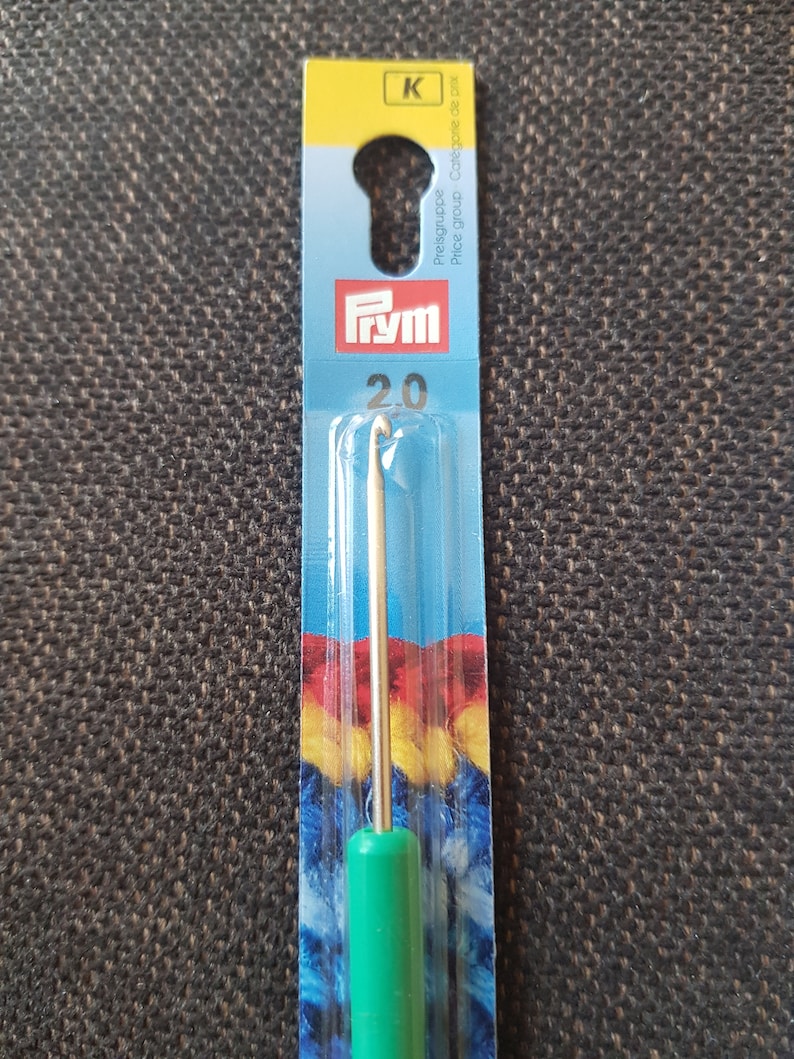 May include: A green crochet hook with a metal hook and a blue and yellow yarn sample on the packaging. The packaging also has the text "Prym" and "20" on it.
