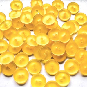 Könnte beinhalten: Eine Ansammlung von durchscheinenden, gelbgoldenen Knöpfen mit gewölbter Form und zwei kleinen Löchern. Die Knöpfe sind auf einer weißen Oberfläche verstreut und erzeugen ein strukturiertes Aussehen. Die Knöpfe sind einheitlich in Größe und Farbe.