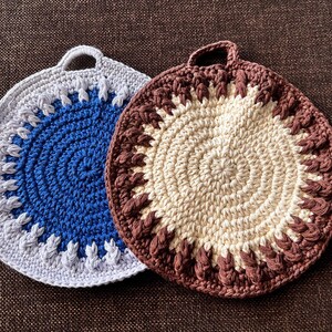 Könnte beinhalten: Zwei handgefertigte Topflappen aus Häkelarbeit. Einer ist blau und grau, der andere cremefarben und braun. Beide sind rund mit einer Schlaufe zum Aufhängen. Das Häkelmuster hat einen strukturierten Rand und eine spiralförmige Mitte.