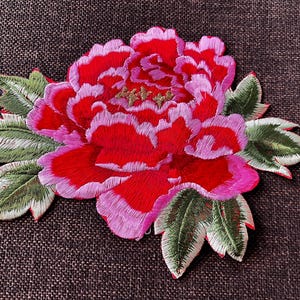 Könnte beinhalten: Gestickter Blumenaufnäher mit einer roten Pfingstrose mit rosa Rändern und grünen Blättern. Die Blütenmitte hat goldfarbene Akzente. Zum Verzieren von Kleidung oder Accessoires.