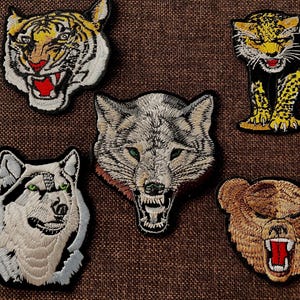 Könnte beinhalten: Gestickte Aufnäher mit Gesichtern verschiedener Wildtiere: ein Tiger, ein Leopard, ein Wolf, ein Husky und ein Bär. Jeder Aufnäher hat detaillierte Stickereien und leuchtende Farben, mit offenem Maul und sichtbaren Zähnen. Die Aufnäher befinden sich auf einem braunen Stoffhintergrund.