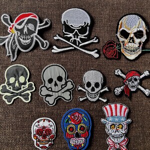 Könnte beinhalten: Eine Sammlung gestickter Totenkopf-Patches in verschiedenen Designs. Einige Schädel zeigen Piratenmotive mit Bandanas und Totenköpfen. Andere beinhalten eine Rose und patriotische Elemente. Die Patches sind in Schwarz, Weiß, Rot und Blau.