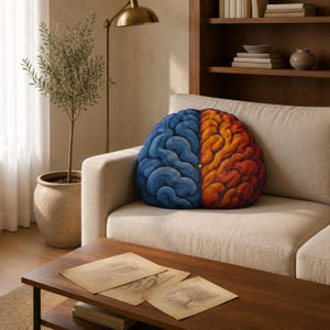 May include: A brain-shaped throw cushion featuring a dual-colour design. The left hemisphere is rendered in shades of blue, while the right hemisphere is depicted in warm tones of orange and red. A small yellow star detail sits at the centre.