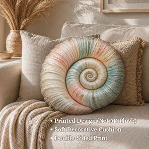May include: A decorative seashell-shaped cushion in pastel colors of pink, blue, and beige rests on a beige sofa. The cushion has a spiral design and is double-sided. The image also shows a beige throw blanket and a textured pillow.