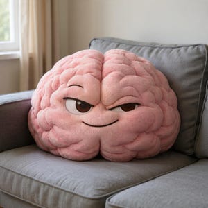 Op de afbeelding: Een roze, hersenvormig kussen met een ondeugende uitdrukking. Het heeft bruine ogen en een sluwe glimlach, liggend op een grijze bank. Het kussen is ontworpen om op een menselijk brein te lijken.