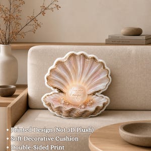 Pode incluir: Almofada decorativa em forma de concha, com um centro em forma de pérola. A almofada é em tons de rosa, bege e dourado, com o texto "You are magic" na pérola. Está sobre um sofá bege.
