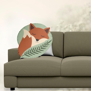 以下が含まれることがあります： 丸い装飾枕。眠っているキツネのイラストが描かれています。キツネはオレンジと白で、緑の葉の上に、薄緑色の背景に寄り添っています。枕は茶色のソファの上に置かれています。