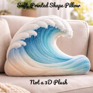 Peut inclure: Un coussin souple imprimé en forme de vague océanique. Le coussin présente des nuances de bleu, de blanc et de beige, avec le texte "Soft, Printed Shape Pillow" et "Not a 3D Plush". Il est posé sur un canapé beige.