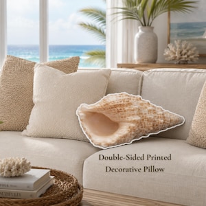 May include: A decorative pillow shaped like a seashell with a tan and cream textured pattern. The pillow features a realistic print of a conch shell. The text Double-Sided Printed Decorative Pillow is visible on the light-colored sofa cushion.