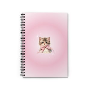 May include: Spiral-bound notebook with a pink cover featuring a kitten illustration. The kitten has gray and white fur and a pink bow. The background is a gradient of pink with star accents. The notebook is designed for writing.