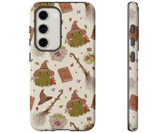 May include: A phone case with a whimsical pattern featuring green frogs wearing witch hats, along with spell books, potion bottles, and brooms. The case has a cream-colored background with a repeating design. The side of the case also has a repeating pattern.