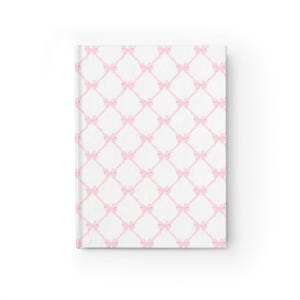 Puede incluir: Una cubierta de cuaderno blanca con un patrón de lazos rosas. Los lazos están dispuestos en un patrón de diamante.