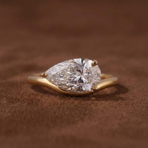 May include: A pear-shaped diamond ring with a gold band. The diamond is clear and faceted, reflecting light. The gold band curves gracefully around the diamond, creating a modern and elegant design. The ring is set against a brown background.