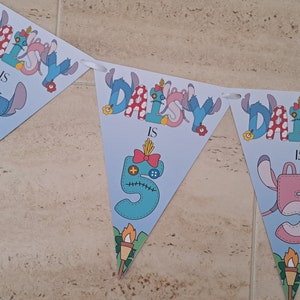 Op de afbeelding: Een blauwe en roze banner met de tekst "Daisy is 5" in een gestikte stijl. De banner is versierd met bloemen en een cartoonfiguur.