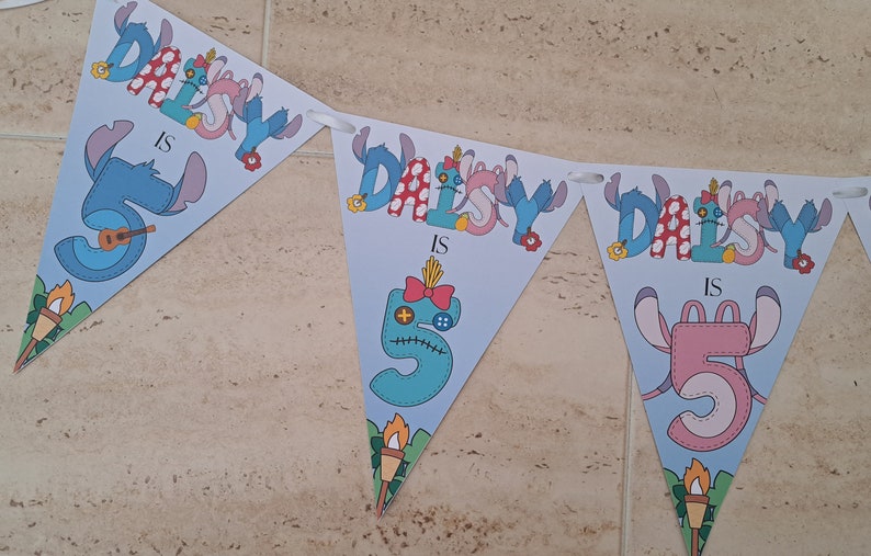 Op de afbeelding: Een blauwe en roze banner met de tekst "Daisy is 5" in een gestikte stijl. De banner is versierd met bloemen en een cartoonfiguur.