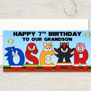 May include: A rectangular birthday banner with a light blue background and the text HAPPY 7TH BIRTHDAY TO OUR GRANDSON OSCAR. The name OSCAR is spelled out in large, colorful letters featuring Sonic the Hedgehog characters including Sonic, Knuckles, Tails, Shadow, and Amy Rose, set against a pixelated ground pattern.