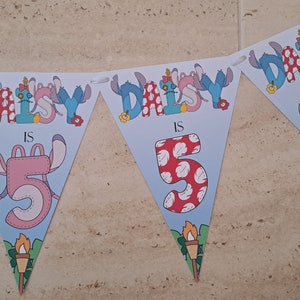 Op de afbeelding: Een blauwe en witte driehoekige banner met de tekst "Daisy is 5" versierd met Stitch-karakters. De banner is gemaakt van papier en heeft een gegolfde rand.