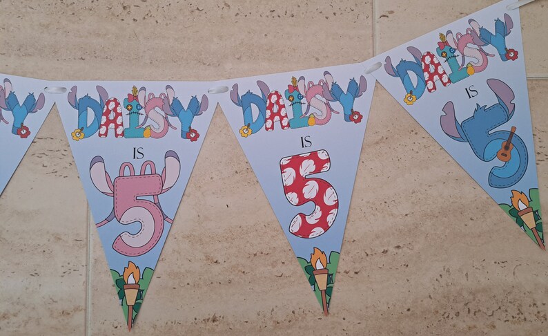 Op de afbeelding: Een blauwe en witte driehoekige banner met de tekst "Daisy is 5" versierd met Stitch-karakters. De banner is gemaakt van papier en heeft een gegolfde rand.