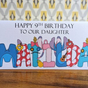 Op de afbeelding: Een verjaardagskaart met de tekst "Happy 9th Birthday to our daughter Matilda" en een kleurrijke illustratie van de naam Matilda gemaakt met gestikte stoffen figuren.