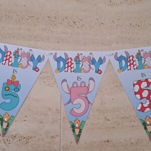 Op de afbeelding: Een blauwe en witte banner met de tekst "Daisy is 5" en een cartoonfiguur. De banner is versierd met bloemen en sterren.
