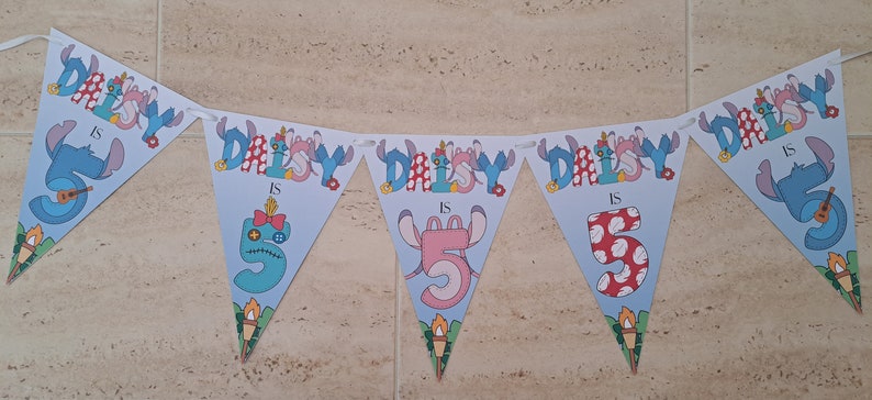 Op de afbeelding: Een blauwe en witte banner met de tekst "Daisy is 5" en een cartoonfiguur. De banner is versierd met bloemen en sterren.