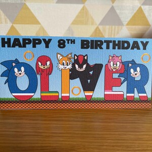 May include: A rectangular birthday card with a light blue background featuring the text HAPPY 8TH BIRTHDAY. The name OLIVER is spelled out in large, colorful letters, each decorated with Sonic the Hedgehog characters including Sonic, Knuckles, Tails, Shadow, and Amy Rose, along with gold rings and a green grass and brown checkered ground pattern.