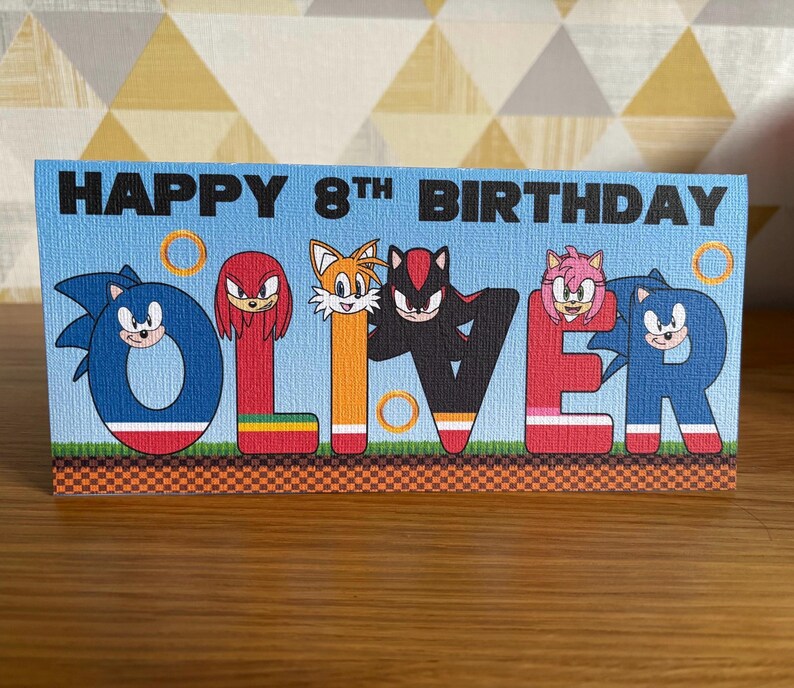 May include: A rectangular birthday card with a light blue background featuring the text HAPPY 8TH BIRTHDAY. The name OLIVER is spelled out in large, colorful letters, each decorated with Sonic the Hedgehog characters including Sonic, Knuckles, Tails, Shadow, and Amy Rose, along with gold rings and a green grass and brown checkered ground pattern.