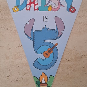 Op de afbeelding: Een blauwe en witte driehoekige banner met de tekst "Daisy is 5" in een gestikte stijl. De letters zijn versierd met een cartoonfiguur met blauwe oren en een ukelele. De banner heeft een tropisch thema met een tiki-fakkel aan de onderkant.