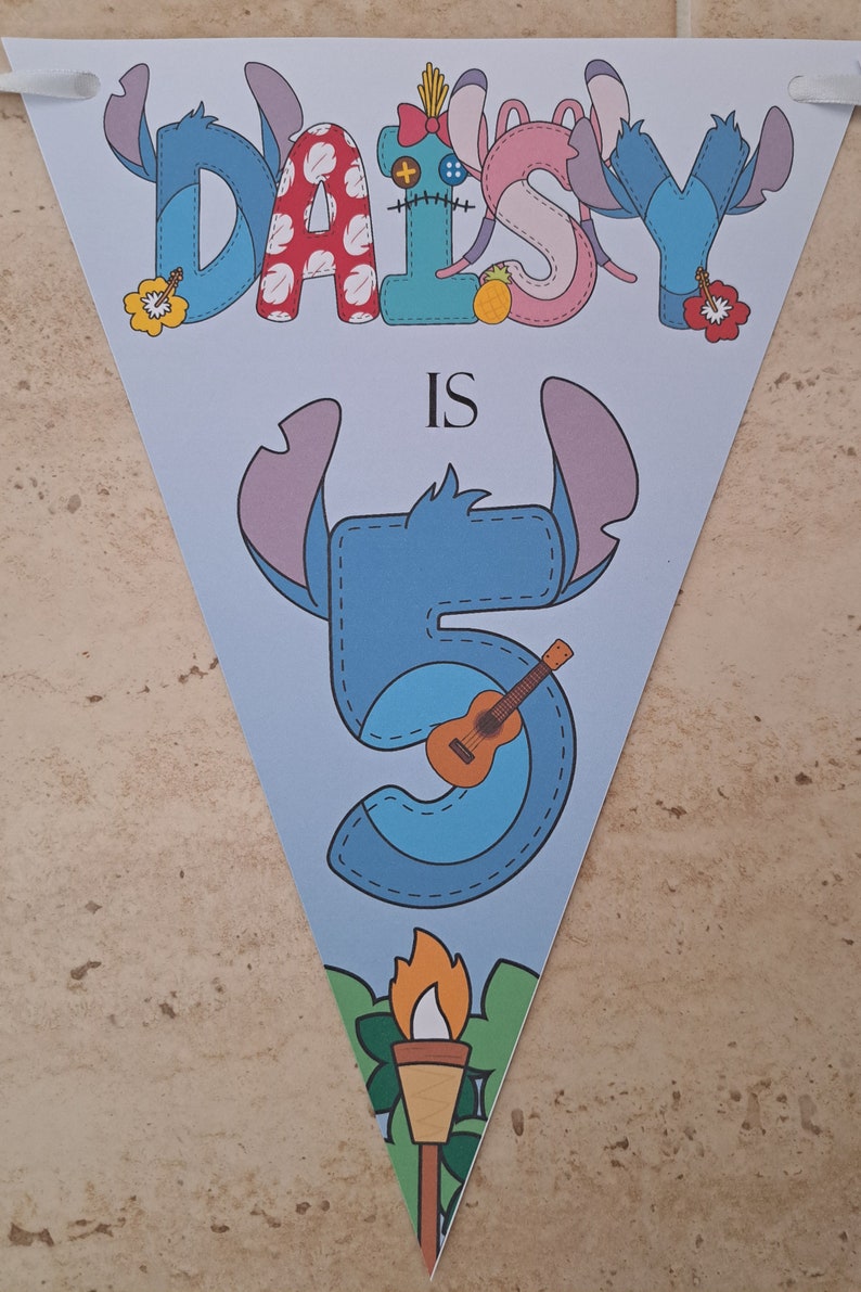 Op de afbeelding: Een blauwe en witte driehoekige banner met de tekst "Daisy is 5" in een gestikte stijl. De letters zijn versierd met een cartoonfiguur met blauwe oren en een ukelele. De banner heeft een tropisch thema met een tiki-fakkel aan de onderkant.