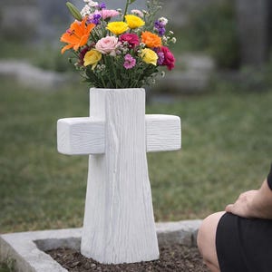 Pode incluir: Um memorial branco em forma de cruz com um vaso cheio de flores coloridas. As flores incluem lírios laranja, rosas amarelas e flores cor-de-rosa. O memorial está num cemitério.