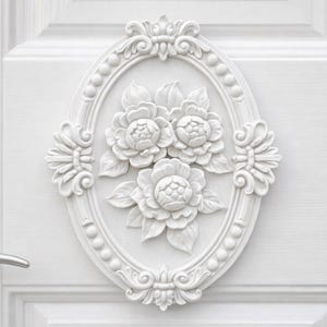 May include: White, ornate floral relief on a white door. The relief features three large flowers within an oval frame, with decorative scrollwork and a silver door handle. The door has a paneled design.