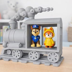 Puede incluir: Un tren de juguete gris con dos figuras de personajes en su interior. Una figura es un perro marrón y canela con uniforme y gorra azules. La otra es un zorro con un impermeable amarillo. El tren tiene una chimenea y ruedas.