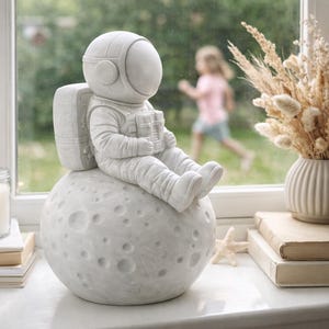 Könnte beinhalten: Eine weiße Astronautenfigur sitzt auf einem strukturierten Mond. Der Astronaut trägt einen Raumanzug mit Helm und Rucksack. Der Mond hat Krater. Die Figur steht auf einer Fensterbank mit einer Vase mit Trockenblumen und Büchern.