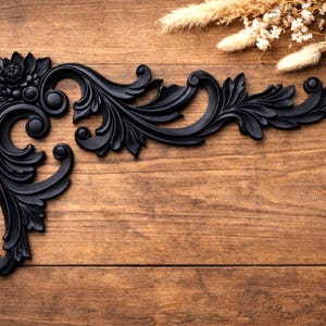 Puede incluir: Moldura de esquina decorativa negra con diseños florales y de volutas. La moldura está colocada sobre un fondo de madera con flores y hierbas secas en la esquina superior derecha. Un elemento decorativo clásico.
