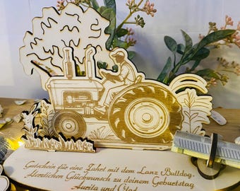 May include: Wooden tractor figurine with a farmer figure, a tree, and a gift card holder. The text on the base reads "Gutschein für eine Fahrt mit dem Lanz Bulldog. Herzlichen Glückwunsch zu deinem Geburtstag Aurita und".
