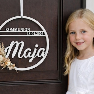 Könnte beinhalten: Weißer Holzkranz mit dem Wort "KOMMUNION", dem Datum "18.04.2026" und dem Namen "Maja". Der Kranz ist mit Trockenblumen und Grün dekoriert. Ein junges Mädchen mit blonden Haaren und einem weißen Kleid steht neben dem Kranz.