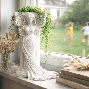 Könnte beinhalten: Eine weiße klassische Statue einer Frau mit fließendem Gewand, die auf einer Fensterbank steht. Die Statue ist mit dekorativen Details verziert und von Grün und Trockenblumen umgeben. Bücher und eine Kerze befinden sich ebenfalls auf der Fensterbank.