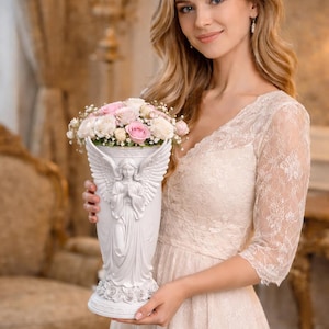 May include: A white angel-themed vase filled with pink and white flowers. The vase has an angel sculpture on the front and is held by a person wearing a white lace dress. The background features a chandelier and ornate decor.