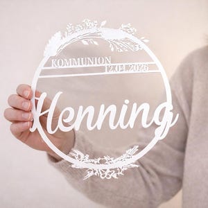 Könnte beinhalten: Ein weißes, kreisförmiges, lasergeschnittenes Schild mit dem Wort "Henning" in Schreibschrift. Das Schild enthält auch das Wort "KOMMUNION" und das Datum "12.04.2026". Florale Designs sind oben und unten im Kreis ausgeschnitten.