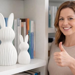 May include: Two white, ribbed ceramic bunny figurines of different sizes sit on a white shelf. The larger bunny is approximately 12 inches tall, and the smaller one is about 6 inches tall. A woman smiles and gives a thumbs up in the background.