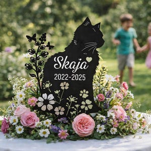 Pode incluir: Placa comemorativa preta em forma de gato com o nome "Skaja" e as datas 2022-2025, rodeada por flores. A placa tem um design de borboleta e floral. Duas crianças estão desfocadas no fundo.