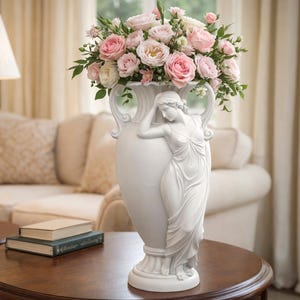 Op de afbeelding: Een witte keramische vaas met een klassiek figuurontwerp, gevuld met roze en witte rozen en groen. De vaas staat op een donkere houten tafel, met een lamp en boeken op de achtergrond. De vaas is ongeveer 61 cm hoog.