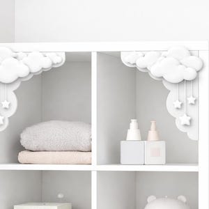 May include: White shelving unit with decorative cloud and star accents. The shelves contain folded towels, two bottles, and a decorative bear-shaped item. The cloud and star decorations are attached to the top of the shelving unit.
