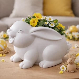 May include: A white ceramic bunny planter filled with yellow and white flowers. The bunny has a smiling face and is surrounded by Easter eggs and a small wooden bunny figurine. The scene is set on a wooden table.