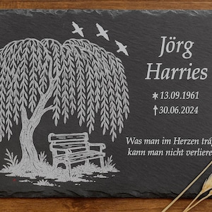 Könnte beinhalten: Schwarze Gedenktafel aus Schiefer mit einer Trauerweide, einer Bank und weißen Vögeln. Die Tafel enthält den Namen "Jörg Harries", Daten und ein deutsches Zitat. Weizenähren sind im Hintergrund.