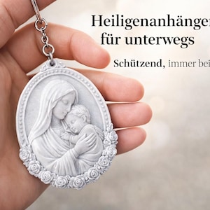 Peut inclure: Un porte-clés pendentif ovale blanc représentant un bas-relief de Marie et l'enfant Jésus, entouré de roses. Le texte sur l'image se lit "Heiligenanhänger für unterwegs" et "Schützend, immer bei Dir."
