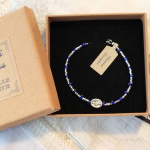 Peut inclure: Un bracelet de perles bleues et argentées, présenté dans une boîte marron avec le texte "Adonai Designs by Hopeville Emporium". Une carte avec une image religieuse est également incluse.
